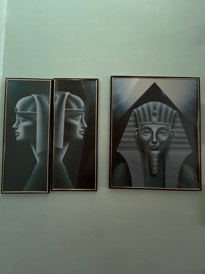 The image displays three framed artworks hanging on a light greenish-gray wall. The photograph itself appears to be rotated 90 degrees clockwise, making the frames appear horizontal in the image, although they would hang vertically on the wall.

The top artwork, if the photograph were correctly oriented, would be the uppermost. It features a grayscale depiction of an ancient Egyptian pharaoh's head, strongly resembling Tutankhamun, wearing a striped nemes headdress with a cobra uraeus. The head faces forward and is oriented vertically within its frame.

Below this, the middle and bottom artworks are also grayscale and depict stylized human profiles. The middle piece shows a smooth, geometric head in profile, facing right within its frame. The bottom piece features a similar stylized head, this time facing left within its frame, possibly an inverted reflection or a complementary design to the middle piece.

All three artworks are enclosed in black frames with thin, gold-colored inner rims. The wall is a solid, untextured color. There is no activity taking place, and no discernible text is visible within the image. The setting is an indoor wall, possibly in Dubovka, Russia, under soft, even lighting.