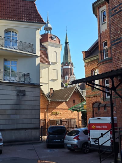 The image shows a street scene in Sopot, Poland, likely in the Old Town or a historic district, with a mix of architectural styles. A pale yellow building with a red tile roof and a balcony with ornate iron railings is on the left. To its right, a section of a cream-colored building with red tile roofs and arched windows is visible, and further back, the green spire of a church or bell tower rises against a clear blue sky. On the right side of the image, a brick building with green decorative accents and arched windows is prominent. In the foreground, a dark grey minivan with a license plate reading "GWE 7050J" is parked, and to its right, a silver hatchback is parked with a license plate "GSP S6XX". A white Poczta Polska (Polish Post) van, a Peugeot Boxer, is also parked in the scene, with the Poczta Polska logo visible on its side. In the middle ground, a small brick building with a green awning is visible, and a sign with "Lotto" can be seen. The overall impression is of a sunny day in a European city with a blend of older and more modern structures.