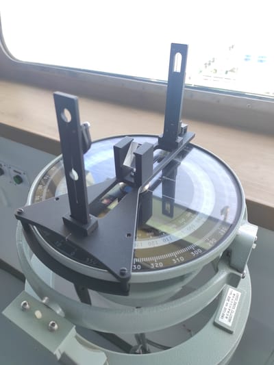 The image displays a close-up of a marine navigational instrument, identified as an azimuth circle or bearing repeater, situated on a ship's bridge or pilothouse. The device is constructed from pale green-grey painted metal and features a large circular glass face. Black metal sighting vanes or arms extend from its center, used for taking precise bearings.

Through the clear glass, multiple concentric scales with degree markings are visible, ranging from 0 to 360 degrees. Prominent numbers such as 000, 010, 020, up to 130, and sequences like 220, 230, up to 350 are clearly displayed in increments of ten. Partial text "Repeater" is also discernible on the instrument's surface.

A label affixed to the instrument's frame indicates "LIHANG Electric Co., Limited" as the manufacturer. Below this, Chinese characters "壁挂式方位分罗经支架" are visible, which translates to "Wall-mounted azimuth compass bracket." The label also specifies "TYPE: B18F" and "S/N: 2511-0543."

The setting is brightly illuminated by natural light entering through a large window to the left, which creates reflections on the instrument's glass. The bright exterior suggests it is daytime. Below the instrument, a part of a control panel with some green buttons is visible. There are no people visible, nor is any specific activity taking place, though the instrument's function is crucial for maritime navigation. The location context for this media file is Kunak, Malaysia.