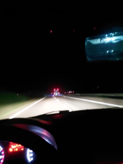 The image depicts a night scene from inside a vehicle. A dimly lit dashboard with red and white indicators is visible. The steering wheel partially obscures the view of a bright roadway illuminated by the headlights of the car. Headlights and taillights of other vehicles are seen in the distance. To the left of the roadway, a dark, blurred object, possibly a bottle, is present.