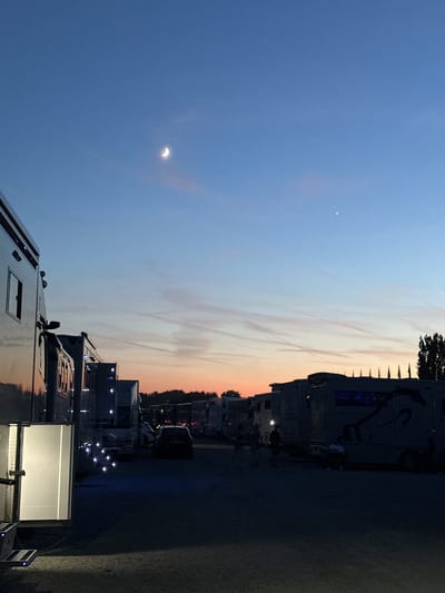 The image captures an outdoor scene at dusk, likely a parking lot or an event venue, with several large trailers or motorhomes parked in a row. The sky displays a gradient of colors, transitioning from a deep blue at the top to shades of orange and pink near the horizon, indicating the sunset. A crescent moon is visible in the upper left quadrant of the sky, accompanied by a faint star.

In the foreground and mid-ground, the vehicles are prominent. The trailer on the far left has its side door open, revealing interior lights and a staircase. Numerous small, square lights are illuminated along the sides of some of the trailers, creating a decorative effect. In the distance, more vehicles and trees are silhouetted against the twilight sky. A dark-colored car is parked among the trailers. A few indistinct figures of people are scattered throughout the scene, some walking and others appearing to be sitting or standing near the vehicles. The ground appears to be a gravel or dirt surface. The overall atmosphere is calm and serene, suggestive of the end of a day's activities. The specific location in Olomouc, Czechia, is not identifiable from the visual elements present.