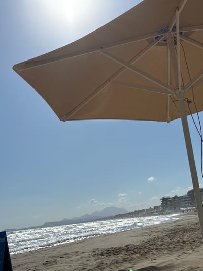 The image captures a sunny day on a beach in Rethymno, Greece. In the foreground, a large, beige beach umbrella with a white frame partially covers the frame, casting a shadow. The umbrella's fabric appears taut, supported by metal struts. Sunlight glints off the ocean waves, creating a shimmering effect as they roll onto the sandy shore. In the middle ground, a line of buildings, likely hotels or apartments, runs along the coastline, with some signs of activity. Distant mountains form a backdrop to the scene. The sky is clear blue with a few scattered white clouds, and the sun is bright, suggesting a warm day. There are no visible people in the immediate foreground, but some figures can be seen on the beach and near the buildings in the distance. A dark blue sign with white text is partially visible on the far left of the frame.