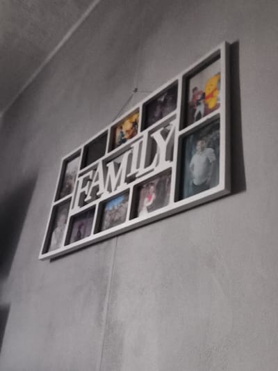 A white multi-photo frame is mounted on a grey textured wall. The frame spells out "FAMILY" with letters in raised relief, interspersed with rectangular and square photo openings. Some of the photo openings contain pictures, including one with a yellow cartoon bear character and two people, and another with a man in a light-colored collared shirt. The frame is tilted, suggesting it is viewed from a low angle. The environment appears to be indoors, likely a residential setting. There is no information in the image that indicates the location is Kohtla-Järve linn, Estonia.