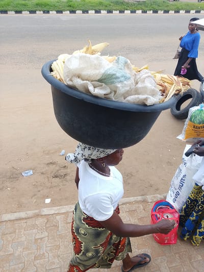 This image depicts a street scene in Oja Oba, Oko Erin, Nigeria. The primary subject is a woman carrying a large black basin filled with corn husks and white sacks on her head. She is wearing a white t-shirt, a colorful patterned skirt, and a headscarf. Her attire and the activity of carrying goods on her head suggest a common practice for vendors in this region.
In the background, another woman in a blue top and black skirt is walking away, carrying a purse and a pink object. The ground is a mix of sand and paved walkway, with some debris scattered around. Several large sacks are visible, one of which has visible text indicating it contains "BANYA RIZ PARBOILED 5%". Another sack has the text "GOOD FRUITS". A red bag filled with colorful straps is also present. The setting appears to be an outdoor market or a roadside area, likely during daylight hours given the brightness of the scene. The weather appears to be fair, with no obvious indicators of rain or extreme conditions. The overall impression is one of daily commerce and activity in a Nigerian community.