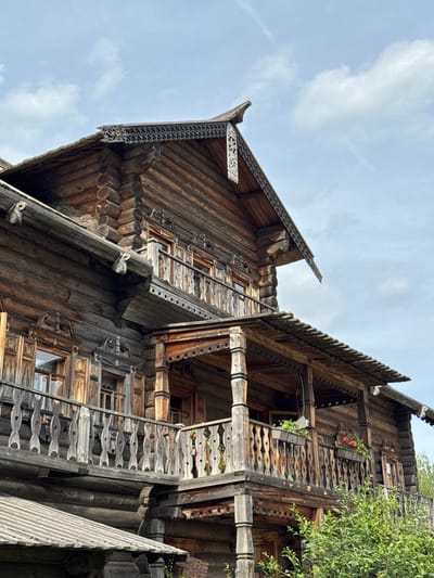 In Nevskii Parkleskhoz, Russia, a close-up, low-angle shot captures a section of a large, historic wooden building constructed from horizontal logs. The building features multiple levels, balconies, and ornate wooden detailing. The dark, weathered wood of the logs shows the texture and age of the structure. On the balconies, intricate wooden railings with decorative cutouts are visible, and window frames are adorned with carved elements. Flower boxes with pink flowers are placed on one of the balconies. The roofline, also wooden, has a decorative, scalloped edge and a triangular gable with carved trim. The sky above is partly cloudy with a light blue hue. Green foliage from bushes or trees encroaches on the lower right side of the frame.