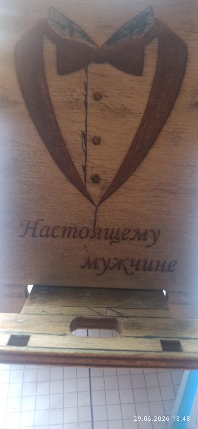 A wooden surface with a laser-engraved design of a tuxedo shirt and bow tie. Below the tuxedo design, there are two lines of text in Cyrillic script. The first line reads "Настоящему" and the second line reads "мужчине". The wooden surface appears to be part of a larger object, possibly a box or a stand, as evidenced by the section of what looks like wooden slats at the bottom of the image. The overall impression is of a decorative item. The image also includes a watermark in the bottom right corner indicating the date "29.06.2026" and time "13:48". The environmental context does not provide any information about the city of Bulaev, Kazakhstan.