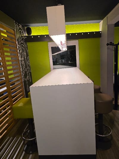 A bright room features a white bar with two round stools, a light fixture, and a decorative wall design. The bar is positioned in front of a green wall with a window. A black wall is partially visible on the left. A wooden screen with slats is at the bottom. The stools have a brown seat and a circular footrest. The overall perspective is a high-angle shot.
