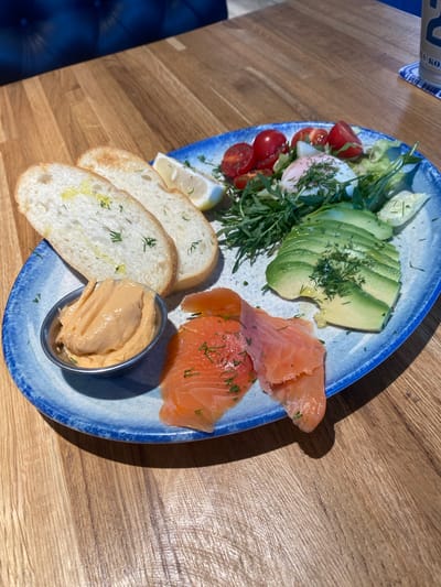 The image shows a close-up view of a breakfast platter on a wooden table. The platter, with a blue rim, contains several food items: three slices of bread topped with oil and herbs, a small metal dish filled with a creamy, orange-colored spread, slices of smoked salmon, sliced avocado, a poached egg on a bed of greens, halved cherry tomatoes, and a lemon wedge. Fresh dill is sprinkled over several of the food items. In the background, a portion of a dark blue upholstered seat and a wooden surface are visible. The setting appears to be a restaurant or cafe. The image is taken from a slightly elevated angle. There are no people visible in the image. The location is identified as Красная Пахра, Russia.