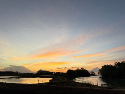This image depicts a serene sunset over a body of water in Grong Grong Capa, Indonesia. The sky is a canvas of soft blues, transitioning to warm oranges and pinks as the sun dips below the horizon. Wispy clouds catch the last rays of light, creating a gentle, ethereal glow. The water, reflecting the colors of the sky, appears calm and still, with dark silhouettes of trees and foliage lining its banks. The foreground is dominated by a dark, earthy landscape, hinting at agricultural fields or marshland. The overall atmosphere is one of tranquility and natural beauty.