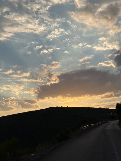 An asphalt road curves uphill along the side of a dark, densely forested hill under a dramatic sky at sunset. The sky is filled with a mixture of light and dark clouds, with streaks of sunlight breaking through, illuminating some of the clouds with warm, golden hues. The top of the hill is silhouetted against the bright sky, with a few sparse trees visible. A metal guardrail runs along the edge of the road. The scene is likely located in or near Višnjevo, Montenegro, given the hilly terrain and vegetation. There are no people or animals visible in the image.