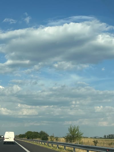 The image shows a road with a white van driving on it, seen from a low angle. The sky is filled with large, fluffy white clouds and a clear blue sky in between. The scene is set in a rural area with a fence and trees lining the right side of the road. The time of day appears to be daytime, with bright sunlight illuminating the clouds. The weather looks pleasant, with a mix of sun and clouds. The van is the primary object in focus, and it is driving on a paved road. The overall impression is a peaceful drive through the countryside.