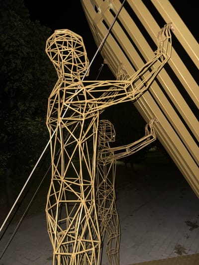 The media file displays a large, wireframe sculpture of a human figure, seemingly in motion, possibly swimming or running. The sculpture is metallic and appears to be gold or brass-toned. It is positioned outdoors, with a dark background suggesting nighttime. Above the sculpture, a structure with horizontal, alternating gold and dark stripes can be seen, possibly an awning or a ceiling with decorative elements. The ground below the sculpture is dark and textured, with hints of what might be paving stones and some foliage visible in the lower left corner. The overall impression is of an art installation in an outdoor setting at night, illuminated by artificial light which casts shadows. The image is taken from a high angle, looking down onto the sculpture. There is no visible text within the frame. The setting appears to be Egorlykskaya, Russia, as provided in the context.