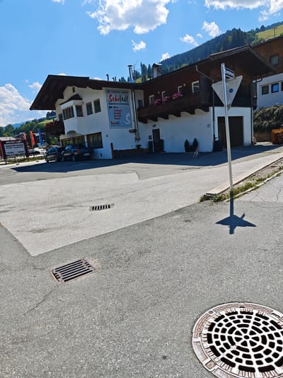 A low-angle, eye-level, exterior shot captures a sunny day in Filzen, Austria, focusing on a building with an attached advertisement for "Schiladl." The building's facade is white with dark wooden eaves and balconies adorned with pink flowers. The advertisement, a large blue and white banner, lists services such as "Verkauf," "Verleih," "Service," and "Depot" alongside the text "Schiladl."
In the foreground, a paved road with a manhole cover is visible. The road leads to the building, where two cars, a dark SUV and a black pickup truck, are parked. To the left of the building, a sign displays information about the "Wagstättbahn" cable car.
Behind the building, a tree-covered hillside rises, culminating in a clear blue sky with scattered white clouds. A triangular street sign indicating "Sonnfeldweg" is also present. The scene suggests a commercial area in a mountainous region.