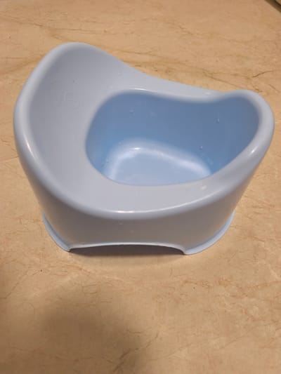 A light blue potty training seat for a child is placed on a beige and brown tiled floor. The potty has a curved backrest and a basin-like seat. There are a few water droplets visible inside the basin and on the outer rim. The floor tiles have a marbled pattern with thin, dark veins. The scene appears to be in a bathroom or similar domestic setting. No people are visible in the image.