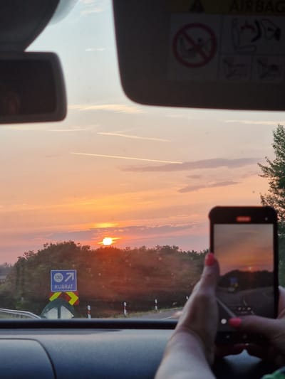 The image captures a sunset scene from inside a vehicle, presumably in Balatonőszöd, Hungary. The viewpoint is from the passenger seat looking out through the windshield. The sun is low on the horizon, casting a warm orange and pink glow across the sky, with faint contrails from an airplane visible.

In the foreground, a person's hand, with bright pink nail polish, is holding up a smartphone, capturing the sunset. The phone screen reflects the scene, showing a blurry image of the sunset and the road ahead.

Outside the vehicle, a road stretches towards the horizon, bordered by trees and vegetation. A blue highway sign with the number "130" and an arrow pointing right is visible, along with text that appears to be "KIJÁRAT," indicating an exit. Red and white striped marker posts line the side of the road.

The interior of the car is partially visible, including the dashboard and a sun visor with safety warning symbols. The overall atmosphere is serene, with the natural beauty of the sunset being documented by the person in the car.
