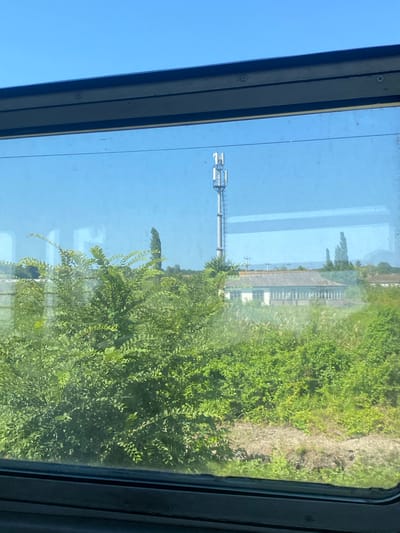 The image depicts a view through a train window, showcasing a rural landscape under a clear blue sky. In the foreground, lush green foliage, likely bushes and trees, partially obscures the view of a low-slung building with a grey roof, suggesting it might be a single-story structure. Behind the building, a tall telecommunications tower with multiple antennas stands prominently, its white structure contrasting with the sky. The overall scene suggests a typical daytime view from a moving train, passing through a sparsely populated area that appears to be in or near the city of Azadogly, Russia. The lighting indicates it is daytime, likely with strong sunlight given the clear sky. There are no people visible in the frame.