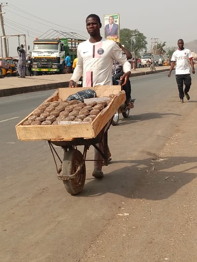 A man in a white long-sleeved shirt with a black Nike swoosh and a "PSG" logo pushes a wooden wheelbarrow filled with numerous brownish, oval-shaped items, likely local food products such as groundnut cakes (kuli-kuli). He has a neutral expression and looks towards the viewer. The scene is set on a sunlit, dusty road in what appears to be a semi-urban or rural area of Nassarawan Eggon, Nigeria, with strong shadows cast on the ground, indicating bright daylight.
In the background, a white Mercedes-Benz truck with a green bed is parked on the left, near other vehicles including a motorcycle and yellow tricycles. Another man in a white t-shirt with "FLIXILIU" and "XL" visible text walks away in the distance on the right side of the road. A large billboard in the upper right corner displays a political figure in a suit with text like "Making a Better..." and "DAVID UMAR". Distant hills are visible on the horizon, completing the landscape of this bustling roadside activity.