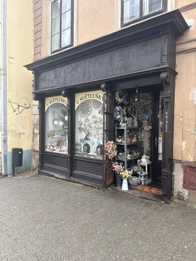 A detailed description of the scene in Znojmo, Czechia, depicts a storefront on a paved urban sidewalk. The building features an ornate, dark-colored facade with two large display windows and an open entrance, identified by arched white signs reading "U SVĚTLUŠKY."

The left display window contains various decorative items, including a large white spherical object, smaller ornate objects, and what appears to be woven or intricate white decorations. The right display window showcases a stylized tree sculpture with colorful elements, multiple circular wall hangings, and other small decorative pieces.

The open doorway reveals an interior densely packed with merchandise. Immediately visible are numerous artificial wreaths, floral arrangements, and a multi-tiered metal shelving unit displaying an array of ceramic animal figures, including hedgehogs and birds. A white ceramic vase filled with artificial flowers (yellow, pink, purple) sits on the ground near the entrance, alongside a white decorative lantern and a stylized metal ornament resembling a gate. A patterned doormat is visible at the threshold. Black graffiti reading "KM-D2016" is present on the right doorframe.

To the left of the storefront, an adjoining building with a faded yellow facade exhibits black graffiti. A black, cylindrical, pole-mounted waste bin is located on the grey paver sidewalk. The building above the storefront features multi-paned windows with dark frames against a faded beige wall. To the right of the storefront, a building with a light brown facade shows a reddish-brown rectangular metal utility access panel. The overall environment suggests an aged city street with wear on the building exteriors. No individuals are present in the image.
