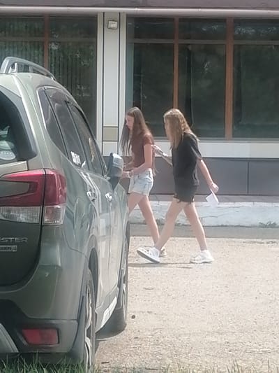 Two young females are walking on a paved surface next to a parked green SUV. The female on the left is wearing a brown t-shirt and light denim shorts. The female on the right is wearing a black t-shirt and dark denim shorts, and is holding a white piece of paper. The SUV is parked on the left side of the image. In the background, there is a building with large windows. The scene appears to be outdoors on a sunny day. The location of this image is the city of Lesozavodsk, Russia.