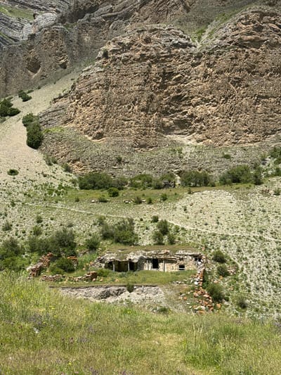 In El'tiubiu, Russia, an ancient stone structure with a grass-covered roof lies nestled in a rocky, mountainous landscape. The structure appears to be a ruin, with broken walls and exposed openings. Green vegetation, including tall grass and scattered bushes, covers the surrounding terrain, with a path leading towards the ruin. The background is dominated by a massive, textured rock face, indicative of a steep mountain slope. The overall scene depicts a remote, historical site within a rugged natural environment.