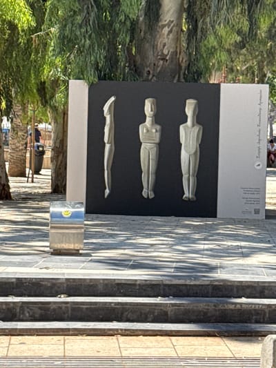 This is an eye-level, medium shot of an outdoor art installation featuring three stylized human figures carved in a light-colored material, set against a black background framed by white panels. The figures are abstract and primitive in design, with distinct heads, torsos, and legs, and appear to be standing side-by-side. The figure on the left is depicted in profile, while the other two are frontal.
To the left of the art installation, a silver, rectangular trash receptacle with a smiley face sticker is visible. The ground in front of the installation is paved with grey stone tiles, and the area is partially covered in dappled sunlight and shadows cast by trees. Behind the installation, lush green foliage and tree trunks suggest a park-like or landscaped area. Further in the background, out of focus, are palm trees, a trash can, and several individuals. One person is standing near the trash can, and others are seated in the distance. The overall scene is bright and appears to be taken during the daytime. The text on the white panel to the right of the artwork appears to be in Greek, possibly identifying the artwork or artist.
The art installation is located at the Archaeological Museum of Heraklion in Heraklion Municipal Unit, Greece. The figures are replicas of the "Phylakopi idols" dating back to the Late Bronze Age.