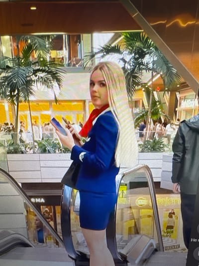 The media file shows a young woman with blonde hair, wearing a blue uniform with a red collar and a white emblem on the sleeve. She is standing on an escalator. In the background, there are large potted plants with palm-like leaves, and a multi-story building with yellow and white elements. There is also a glimpse of a person in a dark grey jacket standing behind the woman, possibly an employee or a companion. The woman is holding a blue object, possibly a phone, in her hand and looking towards the camera with a neutral expression. The scene appears to be indoors, possibly in a shopping mall or a modern public building, given the escalators and the architectural style. The lighting suggests it is daytime. There is some text visible on a sign in the background, which appears to say "HENRY" and possibly "RMA" on another sign. The overall impression is a candid shot of a young woman in uniform in a commercial or public space.