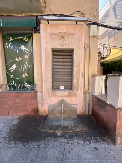 The image shows an outdoor public space in La Garriga, Spain, featuring a water fountain and architectural elements. The fountain itself is made of stone, with water flowing from a spout into a basin. The area around the fountain is paved with dark and grey tiles, with some areas appearing wet from the water. To the left of the fountain, there is a wall constructed of red brick, and above it, a green banner with white dragon-like imagery and yellow leaves is displayed. A security sign for "COVERT SECURITY" is attached to the wall to the left of the fountain. The fountain is situated within a recessed alcove with a pinkish stone facade. Above the alcove, there is a window with horizontal blinds. The wall behind the fountain is also pinkish stone, and there are metal pipes running vertically along it. In the background, a building with a tan facade is visible, with electrical conduits and wires running across it. Further in the background, a yellow awning is visible, along with some greenery and part of another building. The scene is lit by natural sunlight, casting shadows on the pavement.