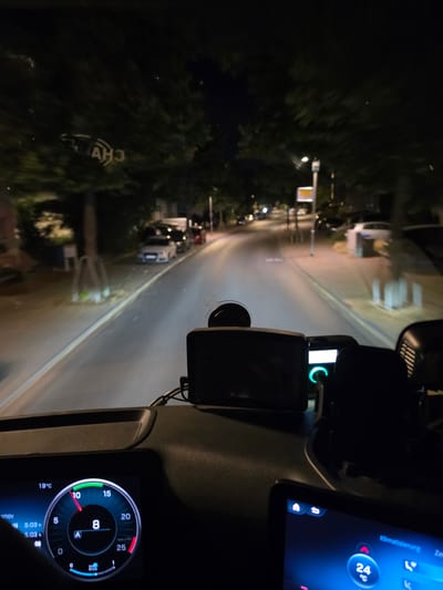 The image captures a nighttime view from inside a vehicle, likely a truck or large van, driving on a street in Flörsheim, Germany. The road is paved, with streetlights and trees lining the sides. Several cars are parked along the street. The vehicle's dashboard is visible, displaying information on a digital screen. A temperature reading of 19°C is shown, along with the time 5:03 h. A circular gauge indicates a reading of '8' and 'A'. Another screen on the dashboard shows "Klimatisierung" and a temperature of 24°C. The overall scene depicts a night drive through a residential or commercial area.