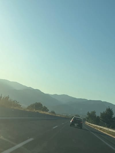 This media file depicts a scenic drive on a highway, likely in Sidirokastro, Greece. The dominant feature is the vast, clear sky, transitioning from a light blue at the top to a pale yellow or white near the horizon. Layered mountain ranges, softened by atmospheric haze, form a dramatic backdrop on the right side of the image. The mountains are densely wooded in the foreground, with sparser vegetation and a lighter green hue further back, indicating distance.
A black car is captured in motion on the highway, positioned in the right lane. Its brake lights are illuminated, suggesting it is slowing down or has recently braked. The highway itself is dark asphalt, marked with white lines delineating the lanes. A white guardrail runs along the side of the road, separating it from the mountainous terrain.
The time of day appears to be either early morning or late afternoon, given the soft, diffused light and the gradient of the sky. The weather seems clear and calm, with no visible clouds. The overall atmosphere evokes a sense of peaceful travel and appreciation for the natural landscape. No people are directly visible besides those potentially inside the car. There is no visible text in the image.