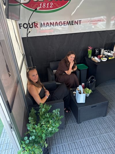 Two women are seated in a covered outdoor area. The woman on the left is wearing a black strapless outfit and sunglasses pushed up on her head, and she is looking towards the camera with a smile. The woman on the right is wearing a brown dress and is looking down while touching her nose. A black coffee table is in front of them with a light blue bag, a potted plant, a can of soda, and cups. Behind them is a black table with a coffee maker, a basket, a vase with pink flowers, and various food items. A banner with the text "FOUR MANAGEMENT" and "CALLIOPE CIVITAVECCHIA" is visible in the background. The floor is made of grey interlocking tiles. The environment appears to be a hospitality or lounge area, possibly at an event. There is no visual information to suggest the location is in Scarperia, Italy.