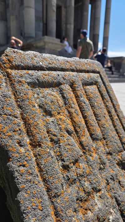 In Garni, Armenia, a close-up shot focuses on a rough, gray stone fragment covered in patches of vibrant orange lichen. The stone features carved, parallel lines running diagonally across its surface. In the blurred background, the classical architecture of the Temple of Garni is visible with its imposing columns. Several people are present, including one individual on the left leaning back with an arm raised, another sitting on steps in white attire, and a person wearing a green shirt and a blue cap facing away from the camera. A person in the background is capturing a photograph. The scene is set outdoors under a clear blue sky.