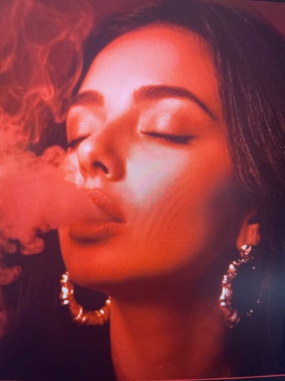 The image is a close-up, medium shot of a woman exhaling smoke, illuminated by a reddish light. Her eyes are closed, and her lips are slightly parted, with smoke wisps around her mouth and extending to the left side of the frame. She is wearing large, hoop earrings. The background is dark and out of focus. There is no discernible indication of the location being Punta Ala, Italy.