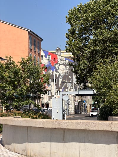 This media file captures a street scene in Charnay-lès-Mâcon, France, bathed in bright, sunny daylight. The view is angled, looking down onto a street lined with buildings and trees.

The foreground is dominated by a large concrete structure on the right, suggesting a bridge or elevated walkway. To the left, lush green trees obscure parts of the scene, with their leaves catching the sunlight.

Behind the trees, a multi-story building is visible, featuring a large mural on its facade. The mural depicts a collection of faces, rendered in black, white, and shades of blue and red. Adjacent to this building are other structures with numerous windows and shutters.

Further down the street, a white car is parked. The street appears to be on a slight incline. Several small, dark objects, possibly birds or insects, are visible against the clear blue sky. The overall impression is one of a quiet urban environment on a pleasant day.

No discernible text is clearly legible in the image.