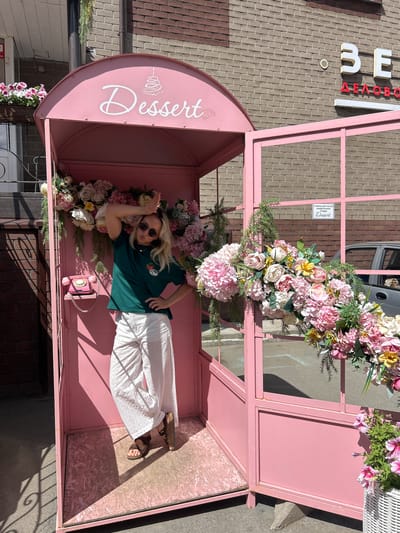 A woman stands inside a pink, phone-booth-like structure adorned with flowers. The structure features a sign that says "Dessert" with a drawing of a cake. A pink rotary phone is mounted on the wall inside. The woman wears a green t-shirt, white pants, sunglasses, and brown sandals. She has her hand on her head and her other hand on her hip. The phone booth is situated in front of a brick building with a sign that reads "BE" in large white letters and "ДЕЛОБО" in red letters. There is a white sign with Cyrillic text and the word "Dessert" visible on the exterior of the structure. The ground is paved, and a car is partially visible in the background. The overall scene suggests an outdoor decorative element for a business, likely a confectionery or dessert shop, in the city of Dulevo, Russia.