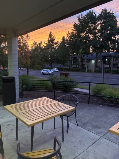 A sunset sky backdrop frames a tranquil street scene. A black metal railing divides the patio, where square tables and chairs sit on a concrete surface. Beyond the railing, a road stretches between lush greenery and buildings. Two cars are parked, their white bodies gleaming faintly. Tall trees silhouette against the orange-hued sky, contributing to the serene evening atmosphere. A portion of the building's roof overhang and a black trash receptacle also appear in the foreground.