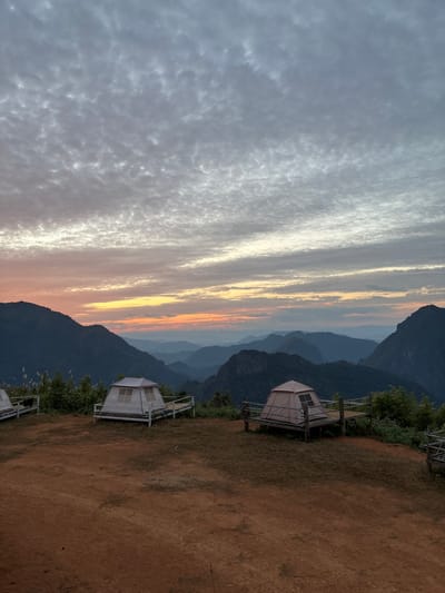 The image depicts a mountain campsite at either sunrise or sunset in Nong Ian, Laos. The foreground consists of reddish-brown earth with sparse grass. Two distinct dome-style tents are visible, positioned on raised wooden platforms. The tent on the left is light grey with white window panels, resting on a white-painted platform with a railing. The tent on the right is tan, featuring a dark window, and is situated on a natural wood-colored platform with a rustic rope and wood railing. Additional platform structures are partially visible on the far left. Beyond the campsite, dark, undulating mountain ranges covered in dense vegetation recede into the distance under a vast sky. The lower sky displays horizontal bands of orange and yellow light at the horizon, transitioning upwards into a lighter blue-grey with widespread, textured altocumulus clouds. No individuals or active interactions are present in the scene.