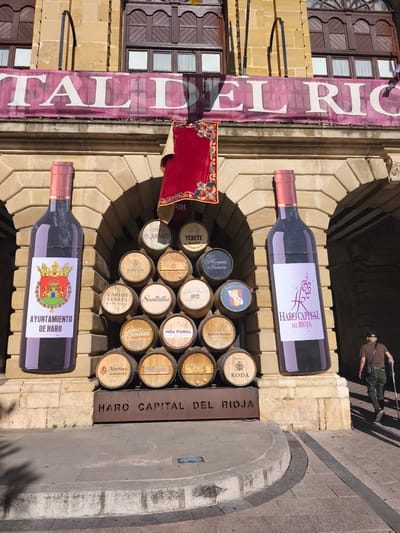 The photograph captures a street scene in the city of Haro, Spain, specifically in front of a building featuring a large banner that reads "LA RIOJA" and below it, "HARO CAPITAL DEL RIOJA." The banner is adorned with ornate decorative elements. In the foreground, to the left, stands a large, upright, two-dimensional cutout of a wine bottle, featuring a label that says "AYUNTAMIENTO DE HARO" and displays a coat of arms. To the right of the bottle cutout, a pyramid of wine barrels is stacked, each barrel bearing various brand names and logos, such as "VIVA OLAVARRI," "TERETE," "Marqués de Carrión," "CARLOS SERRES," "Santalba," "Gomez Cruzado," "VIÑA POMAL," "Martinez Lacuesta," "Muga," "RAMON BILBAO," and "RODA." A smaller brown metal sign is visible at the base of the barrels with the text "HARO CAPITAL DEL RIOJA." Further to the right, a man wearing a brown t-shirt, green pants, and a black cap walks away from the camera, using a walking cane. The building behind the display is constructed of stone and has large arched openings. The ground is paved with concrete slabs and cobblestones. The scene is bathed in sunlight, casting distinct shadows.
