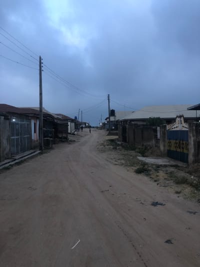 An overcast sky hangs above a dusty, unpaved road winding through a modest neighborhood. Buildings line both sides of the road, ranging from simple structures to a larger industrial-type building with a decorative gate. Utility poles run along the roadside, with visible wires stretching across the sky. The scene appears quiet, with a few distant figures visible further down the road.