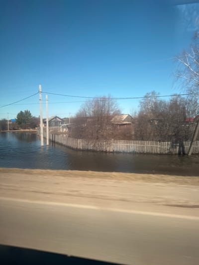The image shows a flooded residential area, presumably in a rural or suburban location. Houses and fences are partially submerged in water. Utility poles are visible, indicating a developed infrastructure. The sky is clear and blue. The photograph appears to have been taken from inside a vehicle. Kueda, Russia is a possibility, but the image's details are not specific enough to confirm this location definitively.
