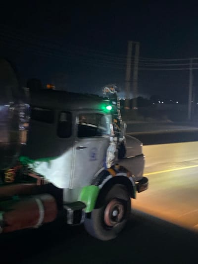 A commercial truck sits alongside a dimly lit road at night. Utility poles and lines appear to the left. The truck’s cab is visible, with a green light illuminated. Various equipment and supplies are mounted on the truck, including what appears to be a large grinding wheel, tanks, and safety gear. The overall scene suggests a roadside repair or utility work in progress. The composition has a somewhat blurred and tilted perspective.