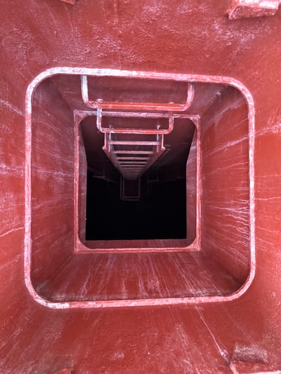This image is a top-down, downward-looking view into a square-shaped metal shaft or opening, likely part of an industrial structure. The interior surfaces are painted a reddish-brown color and show signs of wear, with white streaks and residue, possibly from water or chemical run-off.
A metal ladder is visible, descending into the dark depths of the shaft. The ladder has a handrail on the left side and appears to be made of metal bars. The opening at the top is framed by the thick, reinforced edges of the metal structure, suggesting it is a sturdy, utilitarian design. The overall scene conveys a sense of depth and a potentially enclosed or confined space.
The image does not contain any people, text, or clear indicators of time of day or weather conditions. The location context of Lorca, Spain, does not provide specific visual cues within the image itself, but it places the industrial setting within that geographical region. The activity depicted is a static view into an empty structure, implying it's either a preparation for descent or simply an observation of the space. The overall mood is functional and somewhat stark.