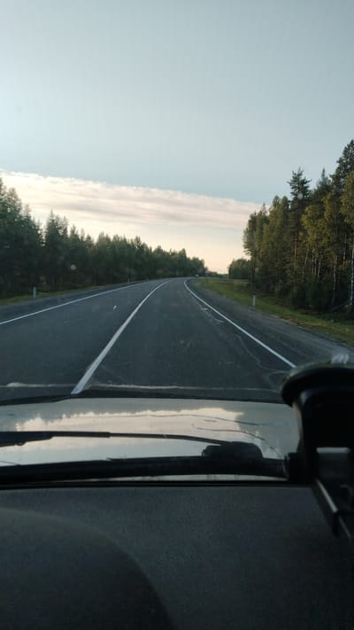 A first-person perspective view from inside a vehicle shows an asphalt road curving through a wooded area. The road has white lane markings. Trees line both sides of the road. The sky is clear with a few wispy clouds. A small dark-colored vehicle is visible in the distance on the right side of the road. The dashboard and windshield wipers of the vehicle are visible in the foreground. The scene appears to be in the city of Vanzozero, Russia.