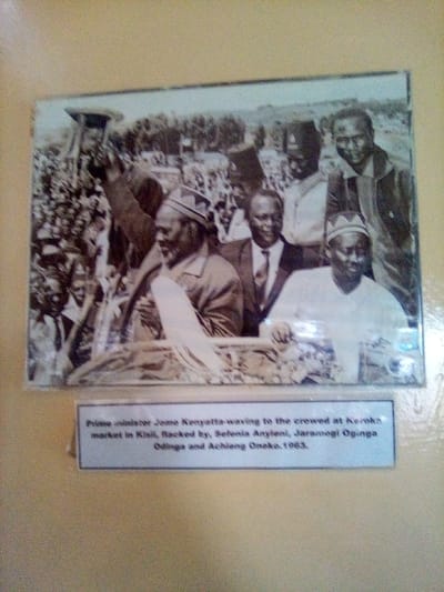 The image, located in South Sakwa ward, Kenya, captures a framed, black-and-white historical photograph mounted on a light beige wall. The photograph depicts a public gathering in Keroka market, Kisii, Kenya, in 1963. In the foreground, Prime Minister Jomo Kenyatta, an older African male with a beard wearing a light-colored jacket and a patterned cap, is prominently featured, mid-wave with his right arm raised while holding a dark object, possibly a hat. To his immediate right, a younger African male in a dark suit and tie smiles. On the far right foreground, another African male in a light shirt and patterned cap looks slightly downwards. These figures are identified by an accompanying white caption card below the framed image as Jomo Kenyatta, flanked by Sefenia Anyieni, Jaramogi Oginga Odinga, and Achieng Oneko. A dense crowd of individuals occupies the mid-ground and background, extending towards a horizon marked by trees and indistinct terrain. The scene suggests the foreground figures are on a raised platform or vehicle, addressing the assembly.