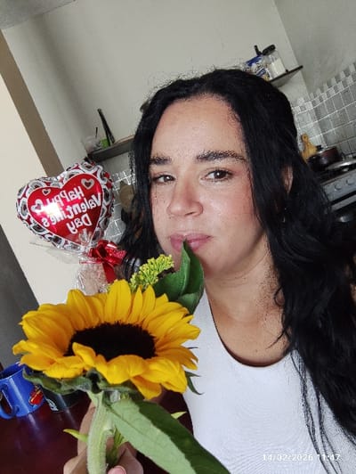A person with long, dark hair and fair skin holds a large sunflower up to their face. The flower's yellow petals are in full bloom, contrasting with its dark center. Behind the sunflower, a heart-shaped balloon with the text "Happy Valentine's Day" is visible. The person is wearing a white shirt. The photo appears to be taken indoors, with a countertop and cabinet visible in the background. The date "14/02/2026" is printed at the bottom of the image.