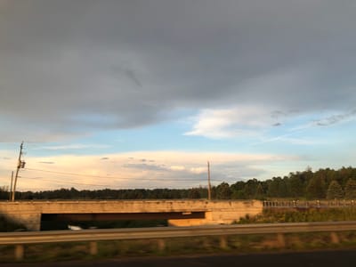 This media file captures a daytime scene of a highway overpass in Innisfil, Canada. The main subjects are the concrete overpass, a body of water visible beneath it, and a dense line of trees forming the background. The sky is a prominent feature, with dramatic, heavy gray clouds dominating the upper portion, transitioning to lighter shades of blue and wispy white clouds closer to the horizon. This suggests a potentially unsettled weather pattern.

The time of day appears to be late afternoon or early evening, as indicated by the warm, golden light reflecting on the clouds and the concrete structure. The warm tones contrast with the cooler blues of the sky. There are no people or animals visible in the frame. The setting is a rural or semi-rural area, evidenced by the abundant trees and the absence of urban development immediately surrounding the overpass.

Notable details include utility poles and power lines running parallel to the road, suggesting this is an active transportation corridor. The bridge itself is a simple concrete structure with a dark metal beam supporting the roadway. A guardrail is visible in the foreground, indicating the vantage point is from a vehicle on a road adjacent to the overpass. A small amount of text is visible on the underside of the overpass, appearing to be a sign, but it is too blurry to be legible. The overall mood is serene, with a hint of atmospheric drama from the clouds.