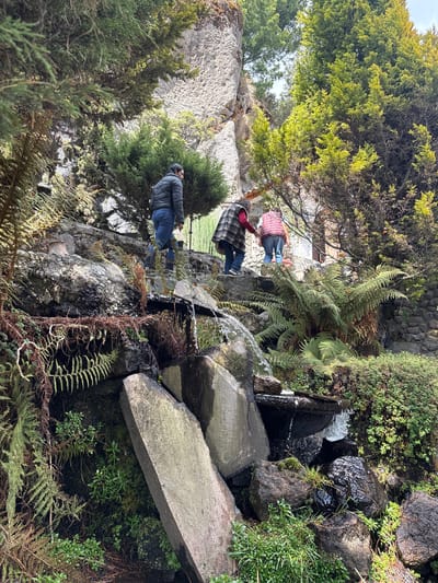 The image shows a landscaped rock garden, possibly a public space, in San Mateo Ozolco, Mexico. Three people are present: a man in a grey jacket, a man in a plaid shirt, and a person in a red shirt. They are walking on a path made of rocks. A small artificial waterfall is present, water flowing over rocks covered in moss and ferns. Various trees and shrubs surround the area. The background shows a partial view of buildings.
