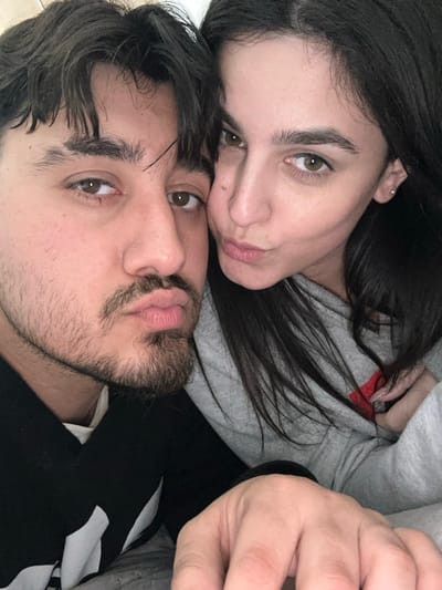 This image, located in the city of Latsia, Cyprus, depicts two individuals in a close-up, upper-body shot. On the left is a male with dark hair and a light beard/stubble on his chin and upper lip. He wears a dark garment with a white element visible on his chest. His lips are pursed. On the right, a female with long, dark hair wears a grey hooded sweatshirt, with a red detail visible on its front. Her left arm is seen resting across the male's chest, with her hand partially visible in the lower right foreground. Her lips are also pursed, mirroring the male's expression. Both individuals possess brown eyes and are looking directly towards the camera. The background is out of focus, suggesting an indoor setting with neutral, light-colored tones.