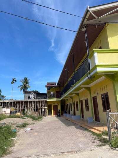 The image shows an exterior view of a yellow building with green trim and a brown roof. The building appears to be an apartment complex or dormitory, with multiple doors visible along the ground floor and a balcony on the second floor. The building is under construction or renovation, with scaffolding visible on one side. In the foreground, there is a paved walkway and some unpaved ground with grass and debris. There are also two palm trees visible in the background, as well as a clear blue sky with some clouds. A woman and a man are visible near one of the entrances, and a motorcycle is parked nearby. The location is identified as Dalam, Indonesia.