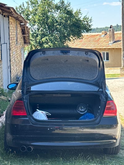 The image shows the open trunk of a black car parked on a grassy area. Inside the trunk, there is a gas cylinder, a white plastic bag, and a blue plastic bag. In the background, there is a brick building on the left, and a house with a tiled roof and a chimney on the right. There are trees and hills in the distance, under a clear blue sky. The scene is set in the city of Konop, Bulgaria.