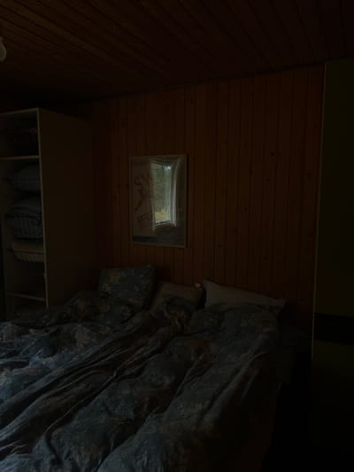 The image shows a bedroom in a house located at latitude 55.558 and longitude 8.157 in Blåvand, Denmark. The room has wood-paneled walls and a bed with a dark blue floral patterned duvet cover.  There is a white framed poster on the wall above the bed that depicts a window looking out onto trees and grass. The bed is unmade with pillows on top. To the left of the bed is a white closet with three shelves, each containing a woven basket. The room is dark and lit only by the natural light coming in through the window.  The image is taken from a slightly elevated angle, looking down at the bed.